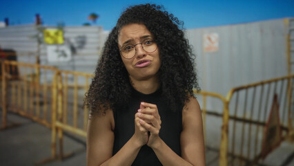 Young woman presses her hands together near a yellow barrier and building entrance outdoors;...