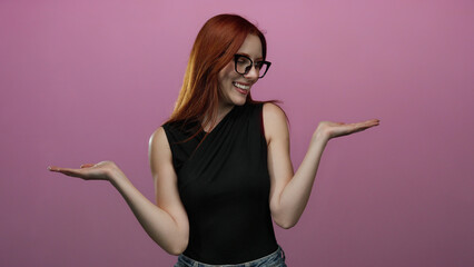Confident woman with red hair wearing glasses stands against an isolated pink background with arms outstretched, exuding beauty and elegance in a stylish black top.