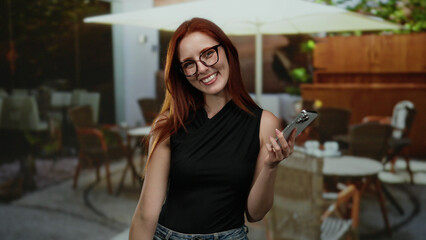 Woman smiling on terrace holds smartphone at outdoor cafe with city vibe around surrounded by...