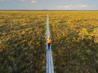 High angle aerial view of a solitary woman walking along a long wooden boardwalk path through a vast, scenic bog.