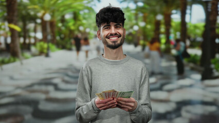 Fototapeta premium Man smiling while holding norwegian krone banknotes on a vibrant urban street with palm trees in the background, showcasing an outdoor financial theme.