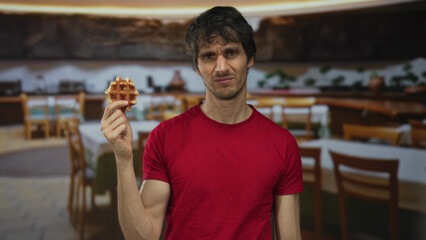 Man in red shirt grimacing at half eaten waffle while standing beside wooden table in restaurant;...