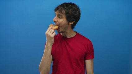 Man in red shirt biting a sugary glazed doughnut with hand to mouth against a blue studio wall;...