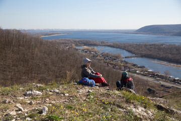 two women  with backpacks relax on a mountain on a hike,  sitting on the grass and enjoy the view  on a sunny spring day.