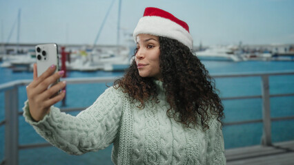 Obraz premium Woman in santa hat taking selfie at seaside port with boats during holiday season, showcasing vibrant outdoor scenery and festive spirit.