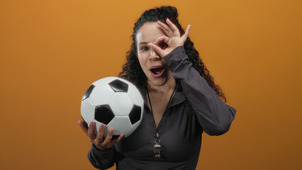 Young hispanic woman making an ok gesture over her eye while holding a soccer ball, standing...