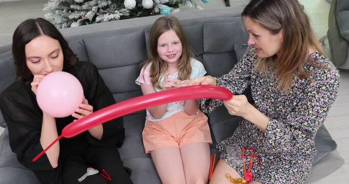 Lesbian women sit close on comfortable couch and blow up colorful balloons for preteen girl. Room begins to fill with floating balloons. Slow motion