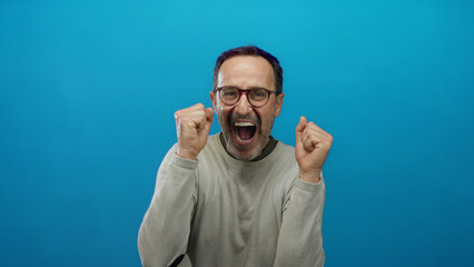 Naklejka premium Mature hispanic man expressing joy and excitement with raised hands against a vibrant blue background, showcasing an enthusiastic and animated expression.