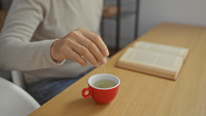 Hispanic man indoors holding teacup while sitting at office desk with open book and dipping teabag showing a calm workplace environment.
