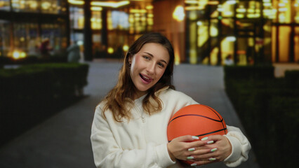 Young woman smiling while holding basketball on city street with illuminated background, enjoying...
