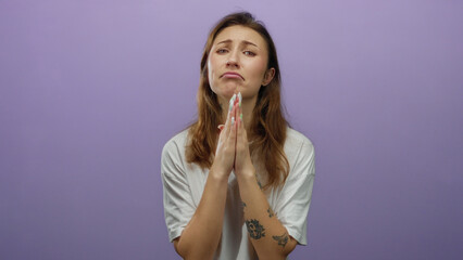 Woman with tattoos in a white shirt pleading with hands together in front of a purple background,...