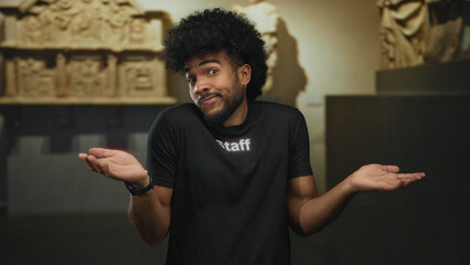 Man with afro shrugging while wearing staff uniform in museum indoor setting, standing against...