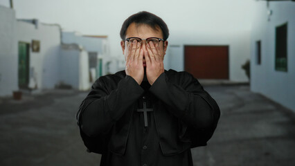 Asian man wearing glasses and black robe with cross stands on an urban street, exuding a sense of contemplation and reflection in a quiet town setting.