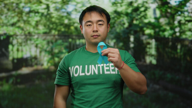 Young man wearing volunteer shirt holds blue cancer ribbon in a park setting showcasing cancer awareness and outdoor participation.