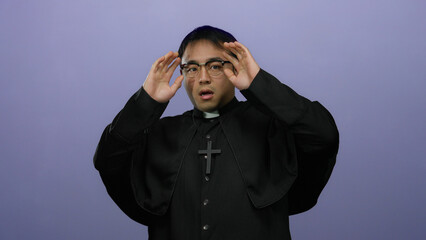 Young man in clerical attire appearing startled against an isolated purple background with raised...