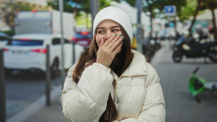 Fototapeta premium Woman smiling warmly in a white coat and beanie on a busy urban street with cars and scooters creating a lively city backdrop under sunny skies.