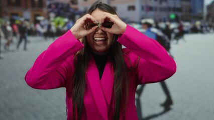 Woman in pink blazer gestures like binoculars with her hands while standing and smiling in a busy...
