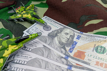 Fanned US dollar bills on a green camouflage background with miniature military jets, illustrating...