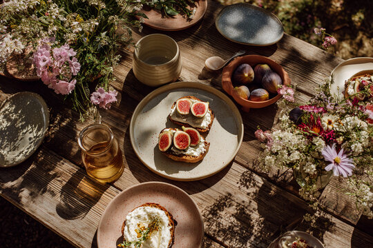 Overhead shot of a rustic wooden table set for a summer brunch with fresh figs, ricotta toast, and wildflower decorations, light shadows falling from surrounding trees