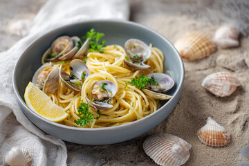 Spaghetti alle vongole served in matte ceramic bowl, garnished with parsley and lemon wedge, surrounded by seashells and a white linen napkin on sandy-textured tabletop