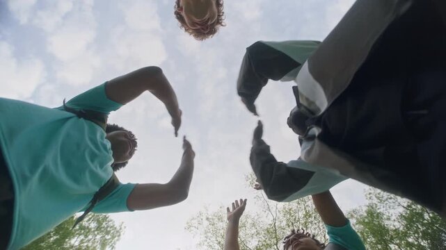 From below shot of multiethnic group of young athletes and their coach exchanging high fives in team circle, celebrating teamwork, success, and mutual support under clear sky