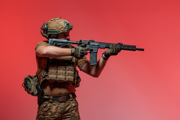 A soldier in military uniform holds a rifle AR 15 in his hands on red background in studio