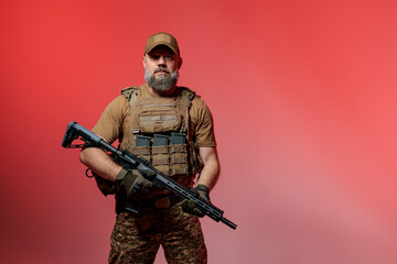 A soldier in military uniform holds a rifle AR 15 in his hands on red background in studio
