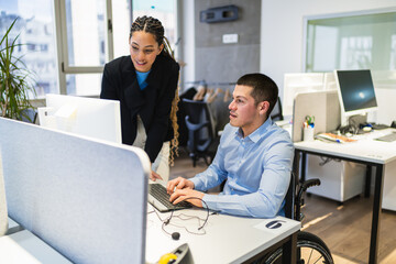 Two young professionals, one using a wheelchair, working together at a computer in a modern...