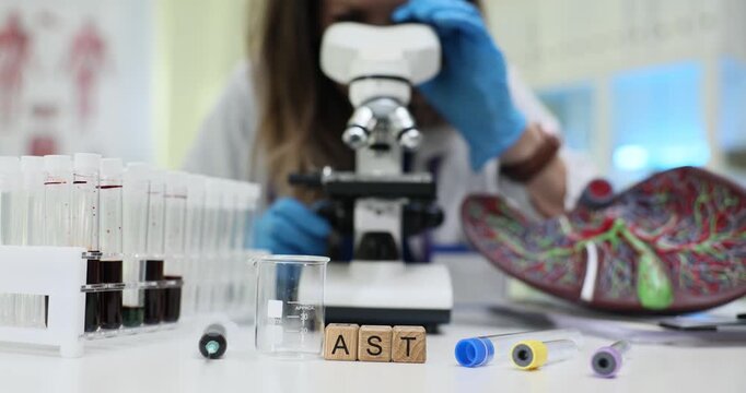 Cubes spell AST standing at microscope and liver model by blood tubes. Word links to enzyme monitoring while lab elements suggest testing in progress