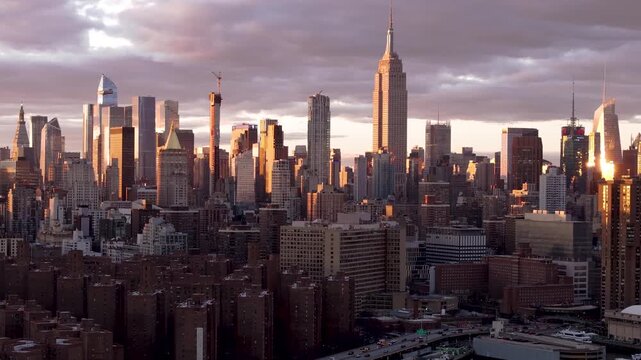 Aerial view of the Empire State Building and Manhattan's skyline bathed in warm sunlight against a backdrop of dramatic, cloudy skies, New York, New York, United States.