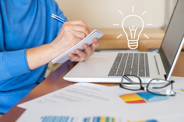 Businesswoman taking notes on notepad while having a new idea, working with laptop and analyzing financial data, a light bulb symbolizes the new idea