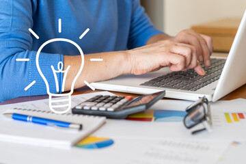 Businesswoman is typing on a laptop with a bright idea light bulb icon, calculator, pen, notebook, eyeglasses and financial charts on a wooden desk, symbolizing innovation and business analysis