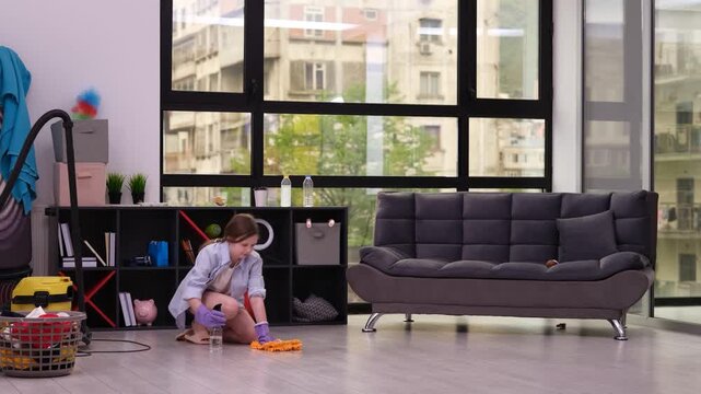 Teen schoolgirl cleans floor using rag kneeling near toy basket and shelves. Arms move back and forth scrubbing surface with visible determination