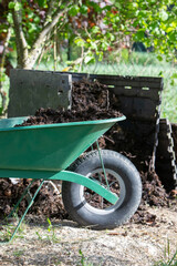 Une brouette verte remplie de compost, scène dans un jardin. Bac de compost ouvert en arrière plan. Promouvoir le recyclage durable et les pratiques de compostage domestique.