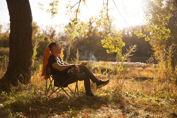 A middle-aged man enjoys smoking a cigar while sitting in an armchair on a sunny autumn day in...