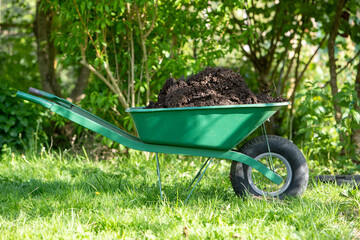 Une brouette verte remplie de compost, scène dans un jardin. Bac de compost ouvert en arrière plan. Promouvoir le recyclage durable et les pratiques de compostage domestique.