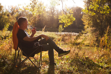 A middle-aged man enjoys smoking a cigar while sitting in an armchair on a sunny autumn day in nature