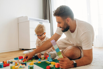 Sitting on the floor. Father is playing in the room with his little son with construction sets
