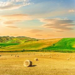 Scenic view at picturesque burning sunset in a green shiny field with hay stacks, bright cloudy sky , golden sun rays and road leading far away, summer valley landscape