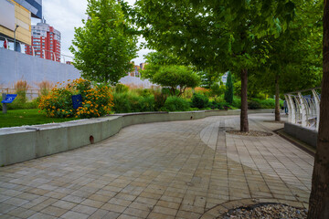 A modern urban park pathway winding through lush green trees, vibrant yellow flowers, and manicured landscaping, with contemporary buildings in the background, offering a peaceful city escape.