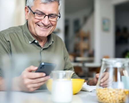 Smiling senior man enjoys breakfast while using smartphone.