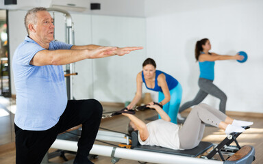 Willing aged man engaging in pilates training on pedal fitness chair in exercise room during workout session. Persons practicing pilates with trainer