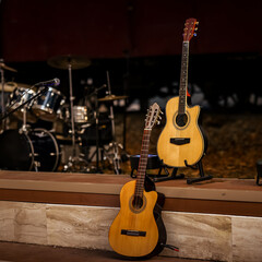 Two acoustic guitars lean against a stage edge in warm light, set before blurred drums and a...