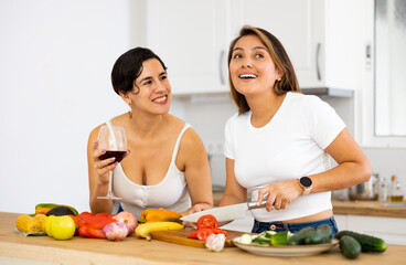 Smiling young Hispanic woman cooking with sister in home kitchen, chatting cheerfully and drinking wine while preparing vegetable salad. Happy family relationship concept..