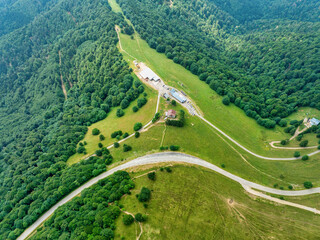 Aerial view of a winding road cutting through verdant hillsides and dense forests, leading to a clearing with buildings, Oderen, Grand Est, France.