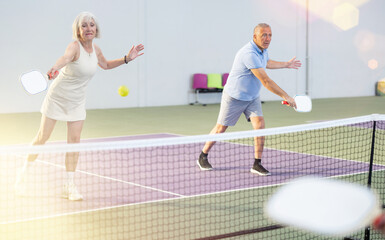 Concentrated experienced team of pickleball players, aged woman and man, preparing to strike and return ball to opponents field during doubles match on indoor court