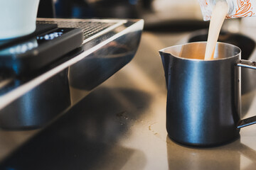 Close-up of milk being poured from a carton into a metal frothing pitcher in a coffee shop setting,...