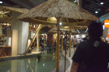 A blurred photo of a tropical-themed mall interior with thatched umbrellas, a water feature, and indistinct shoppers, creating a serene yet lively commercial atmosphere.
