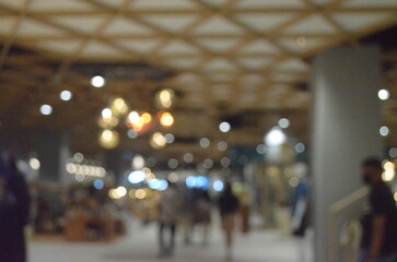 A dreamy blurred photo of a mall interior, showcasing warm glowing lights, a wooden ceiling, and faint silhouettes of people, evoking a modern urban shopping atmosphere.
