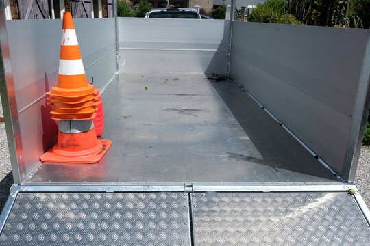 Aluminum trailer with ramp on gravel driveway near cozy house and vibrant garden under sunny, cloud-speckled sky. Loading platform.